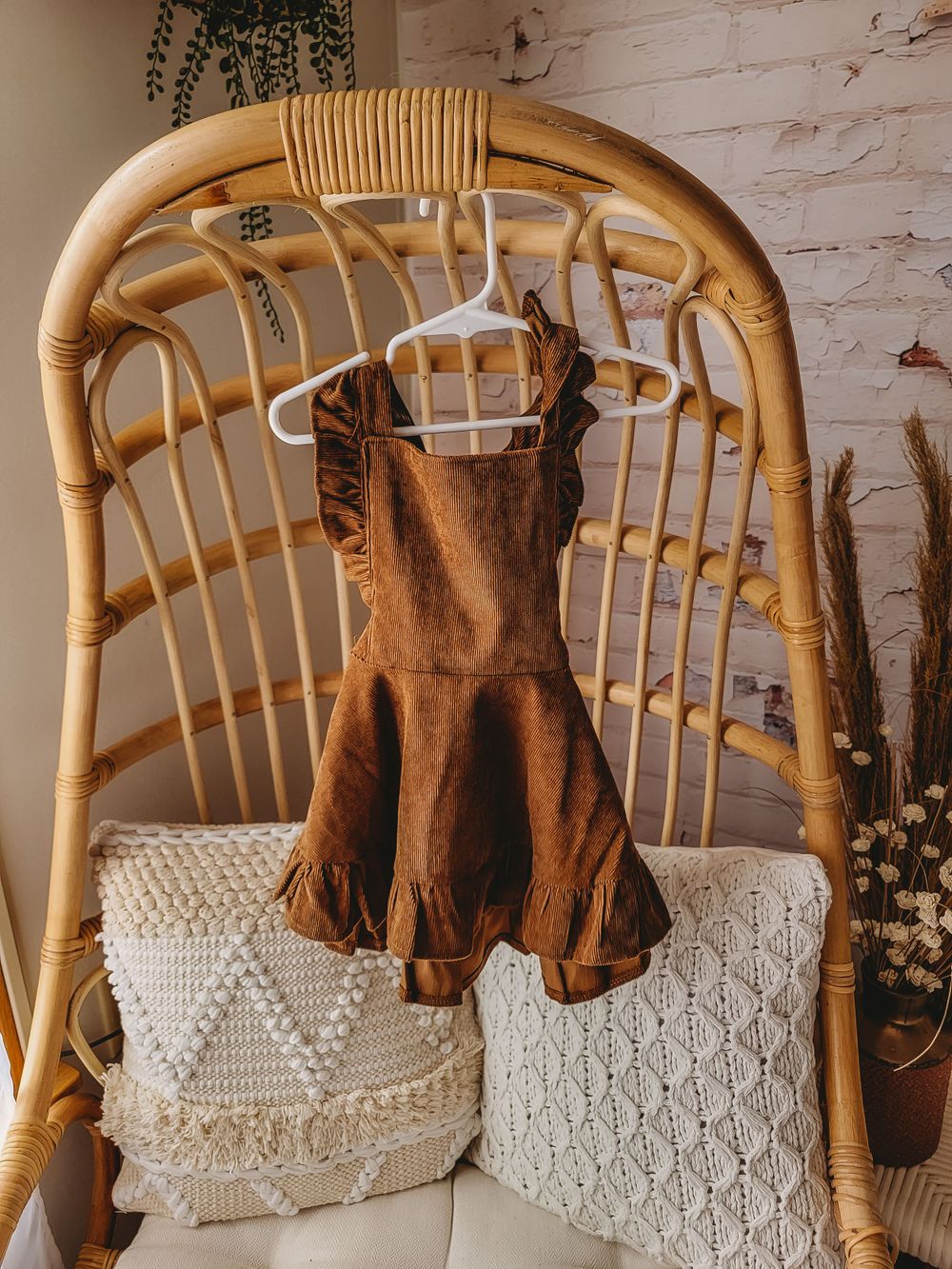 Brown ruffle smocked toddler dress on white hanger displayed on rattan chair with boho pillows.