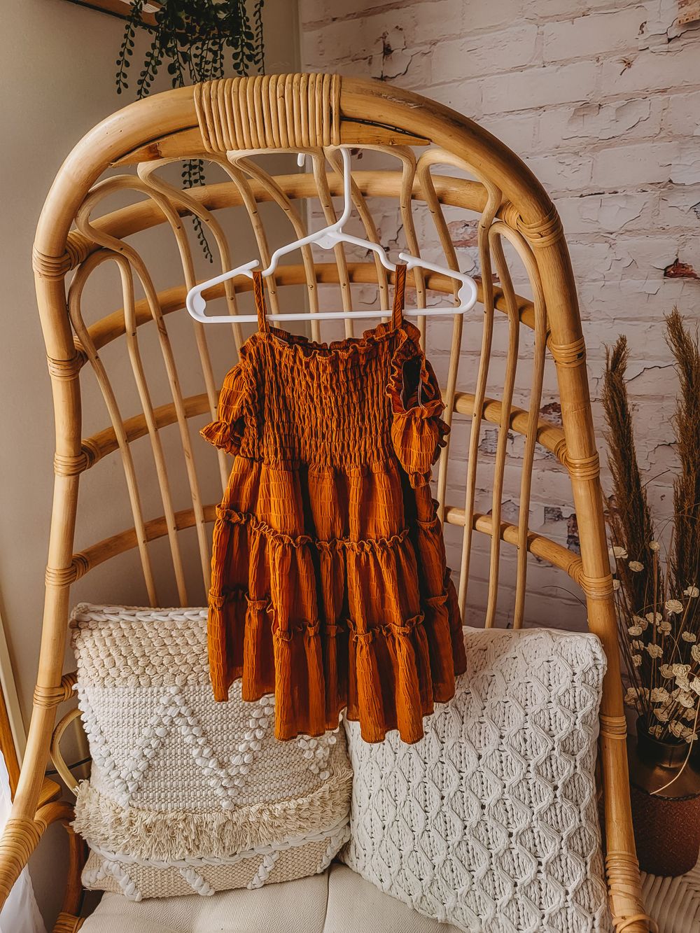 Rust orange tiered smocked toddler dress on hanger displayed on rattan chair with neutral pillows.