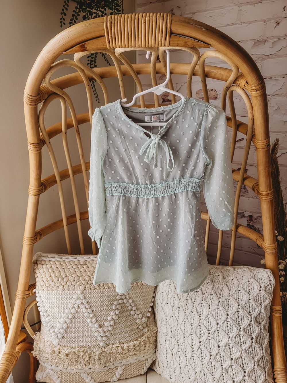 Sage green dotted swiss long sleeve baby girl dress with tie neck on rattan chair.