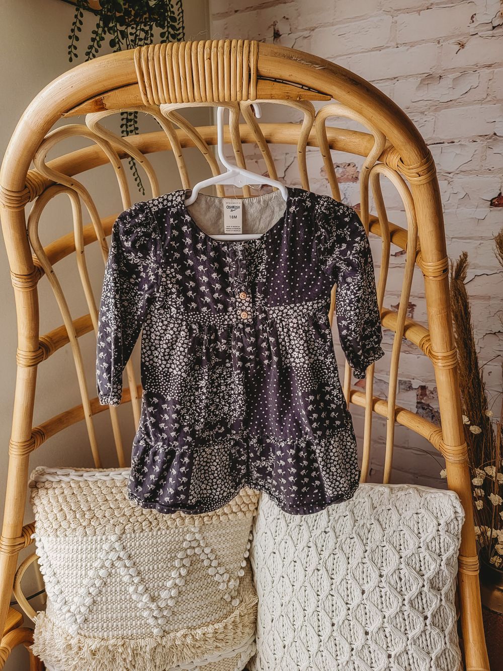 Dark floral print long sleeve tiered baby girl dress on hanger on rattan chair.