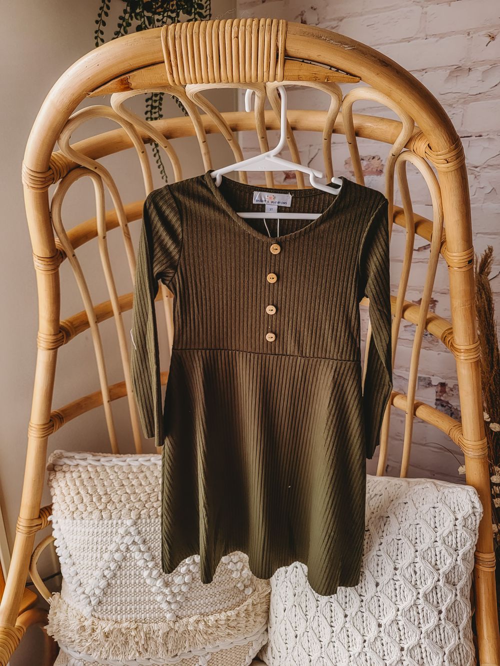 Olive green ribbed long sleeve henley button toddler dress on hanger on rattan chair.