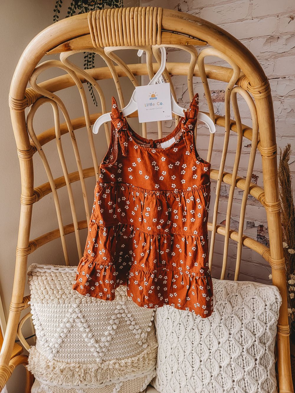 Rust orange floral sleeveless tiered baby dress on a white hanger displayed on a rattan chair.