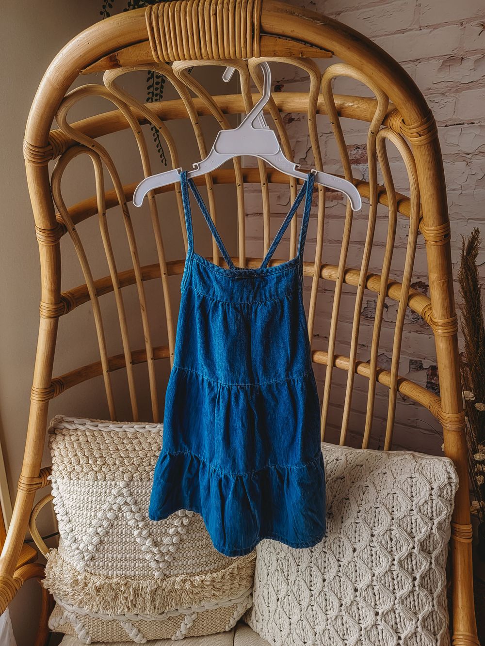 Blue denim spaghetti strap tiered toddler sundress on white hanger on rattan chair.