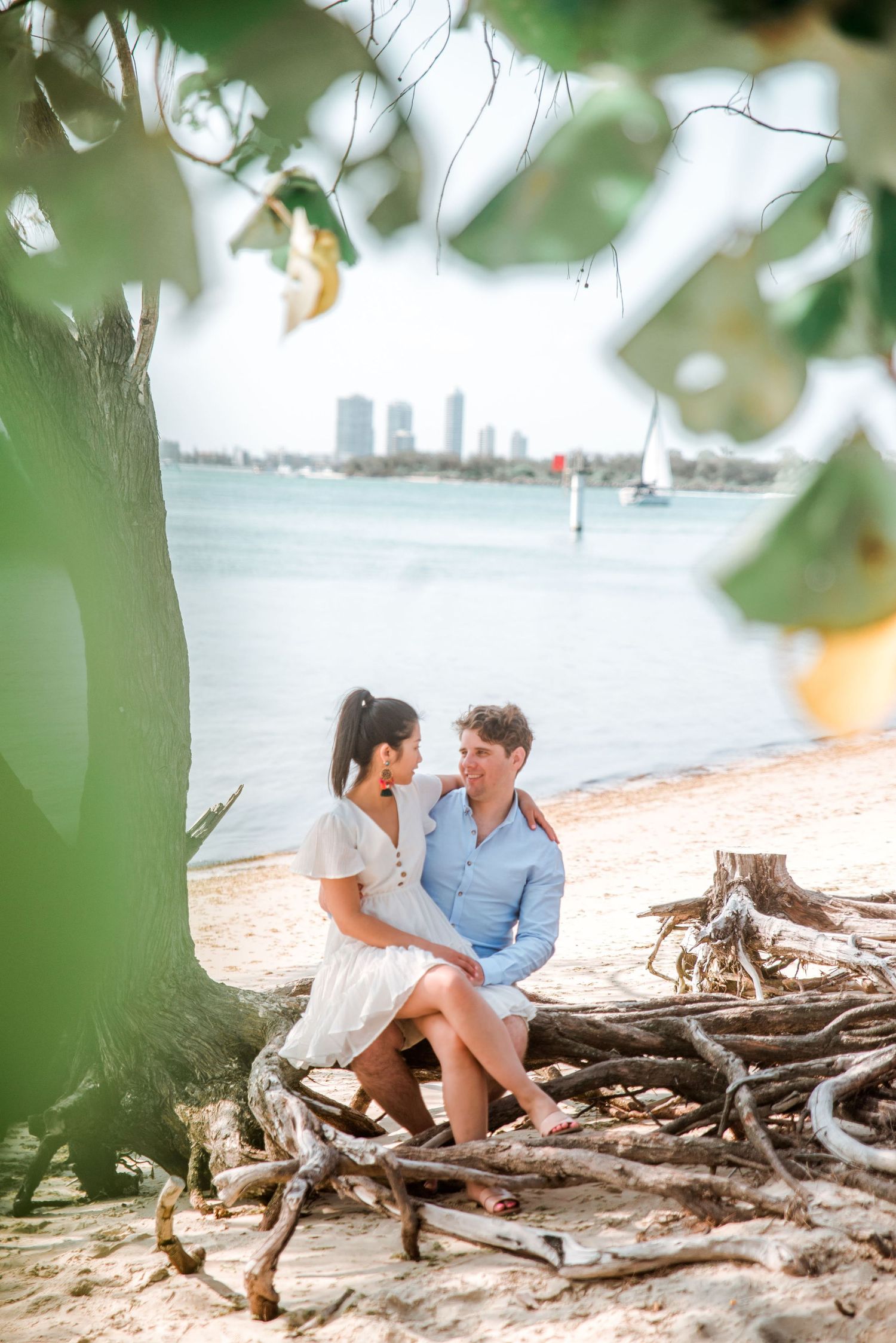 A couple shares an intimate moment sitting on driftwood by the water with cityscape in background.