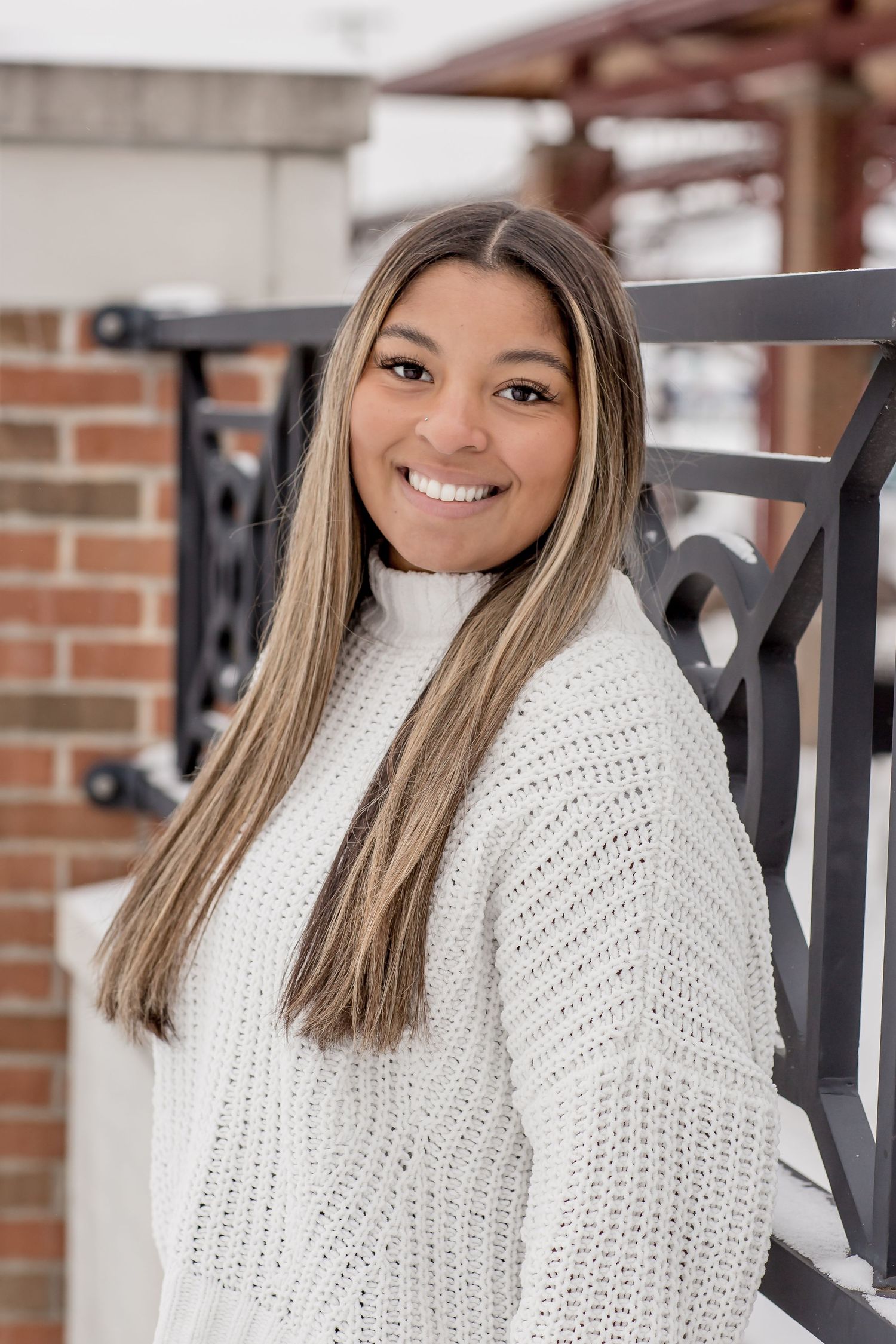 Smiling young woman with highlighted hair wearing a white knit sweater leaning against iron railing.