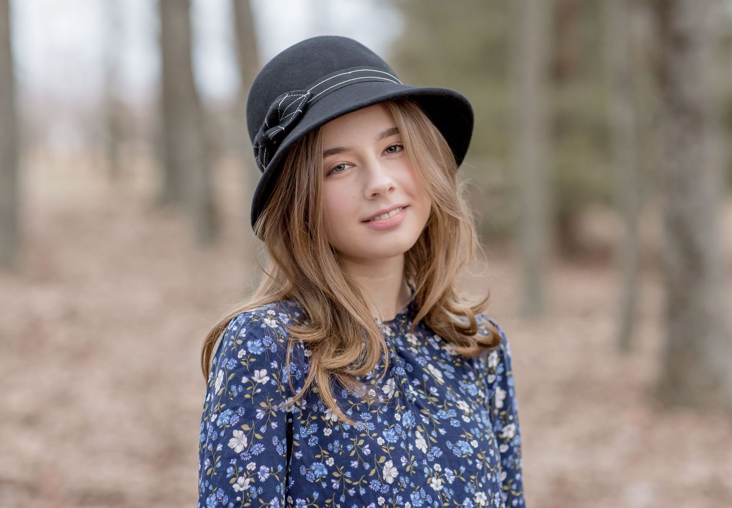 Young woman wearing a black cloche hat and floral blue dress posing outdoors in a forest setting.