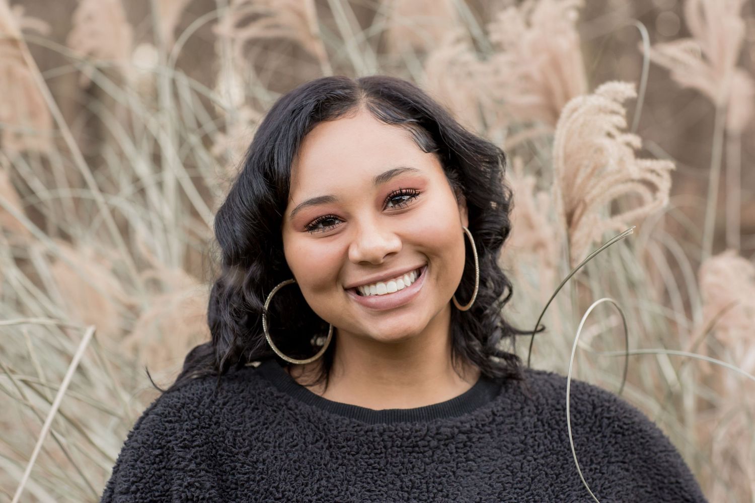 Smiling young woman with curly black hair in black top poses among tall dried grasses.