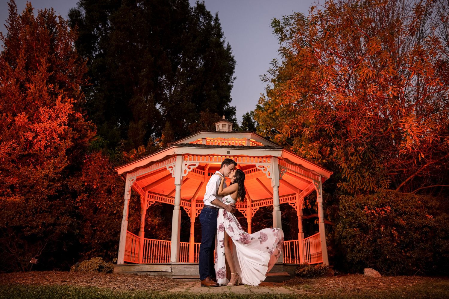 Newlyweds pose dramatically in red-lit gazebo surrounded by vibrant autumn foliage at dusk.