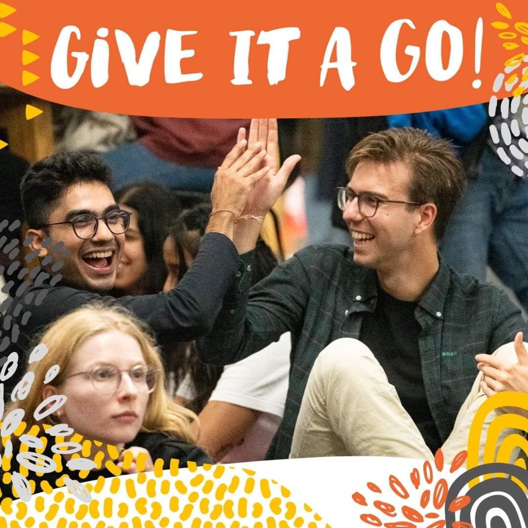 A group of friends celebrate enthusiastically under an orange 'Give it a go' banner.