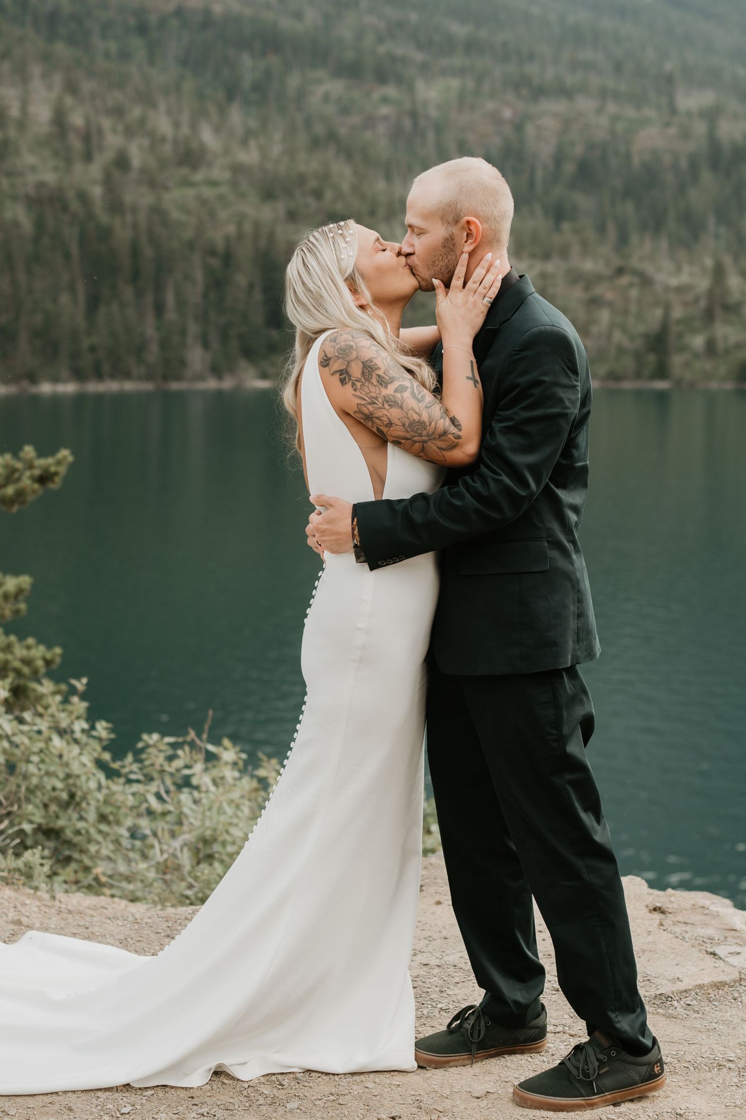 Intimate wedding kiss captured against mountain lake backdrop.