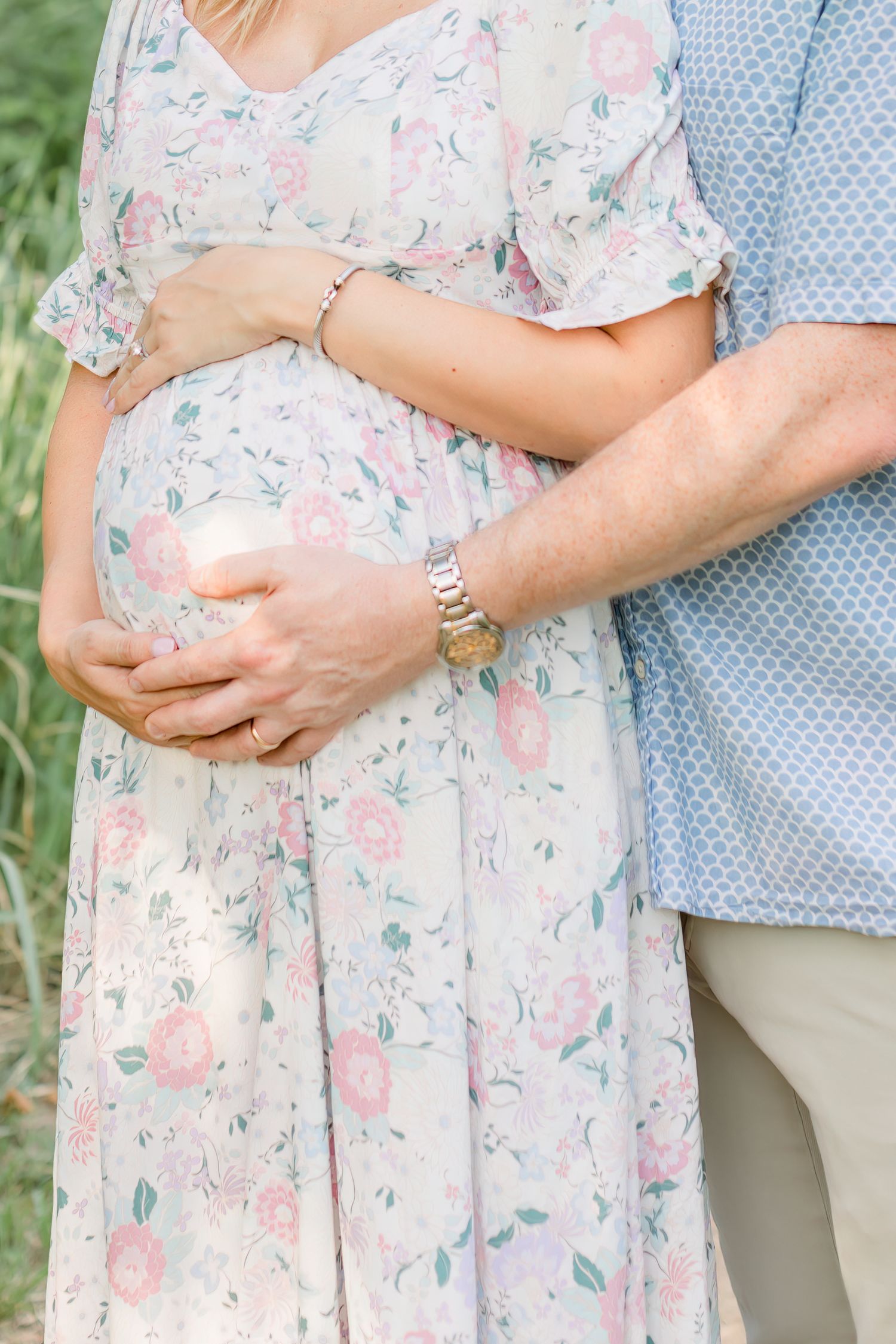 Close-up of hands cradling a pregnant belly in a floral dress against natural scenery.