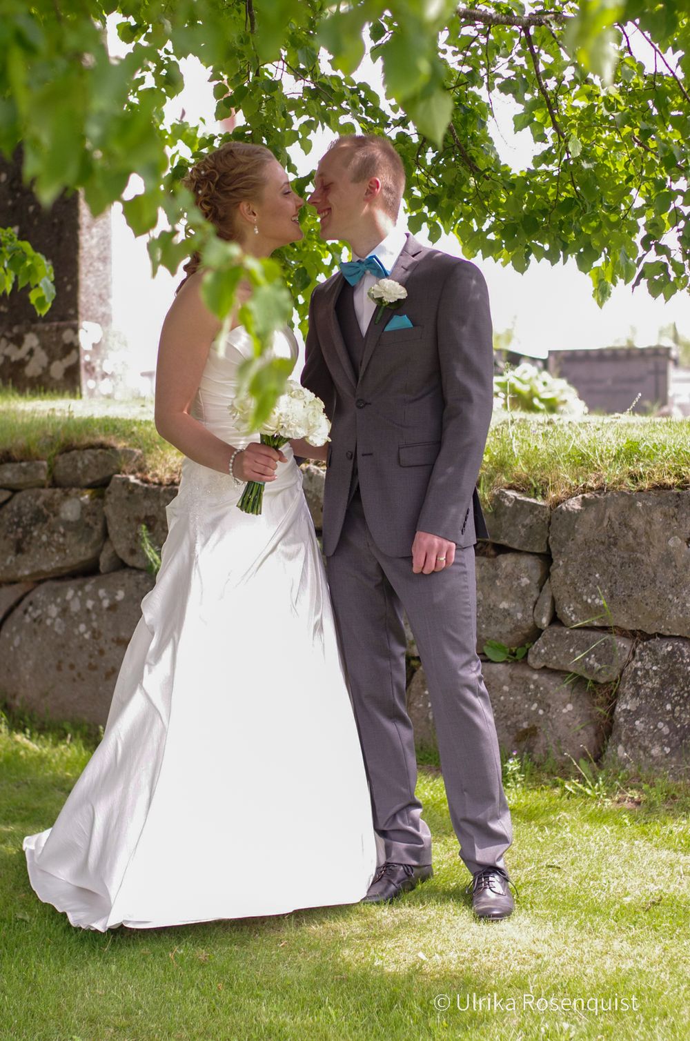 A romantic wedding couple embracing under green leaves near a stone wall.