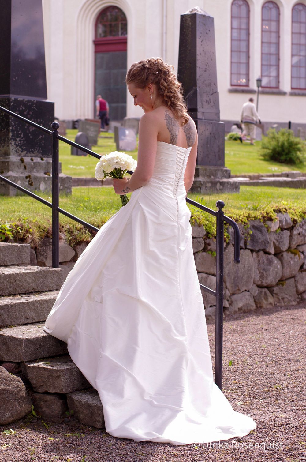 A strapless white wedding dress with a bouquet photographed on stone steps by a church.