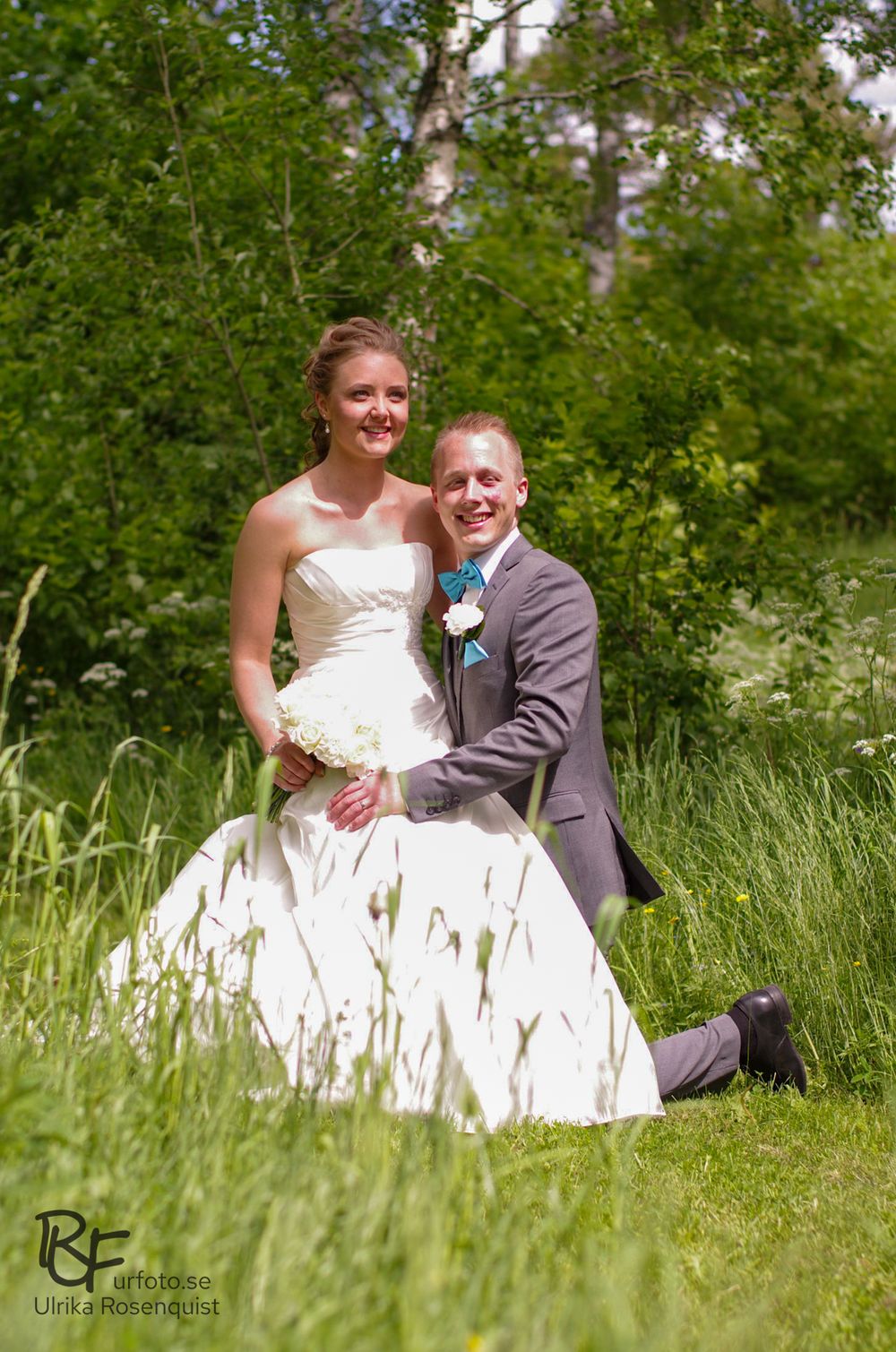A wedding couple posing in tall summer grass with lush greenery background.
