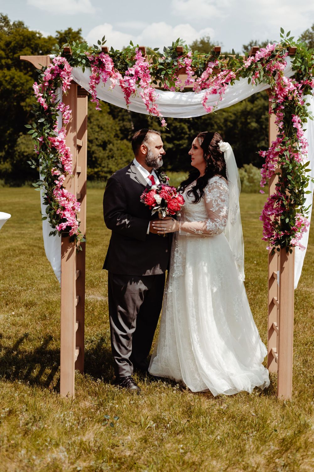 An outdoor wedding ceremony under an arch decorated with pink flowers and white fabric.