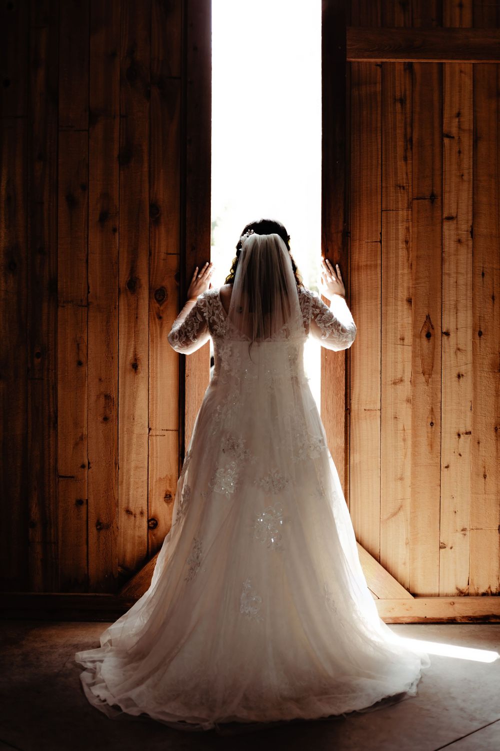 A dramatic backlit silhouette of a bride in a lace wedding gown standing in front of wooden doors.