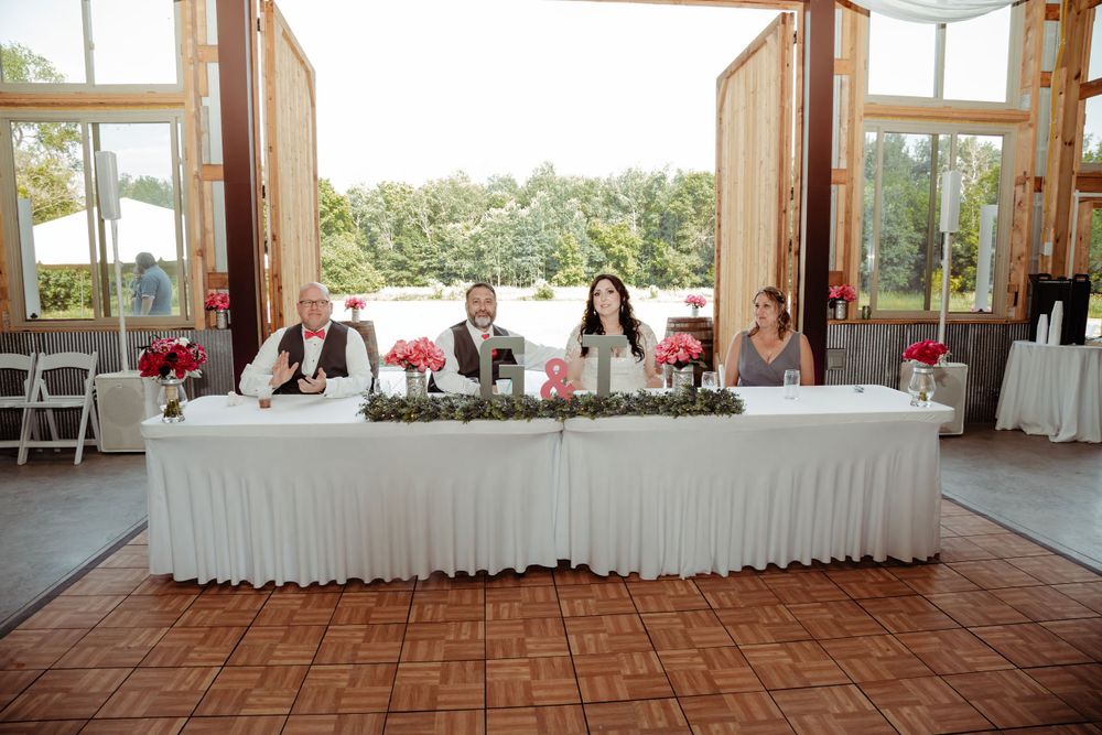 Wedding reception head table setup in a rustic barn venue with white linens and red accents.