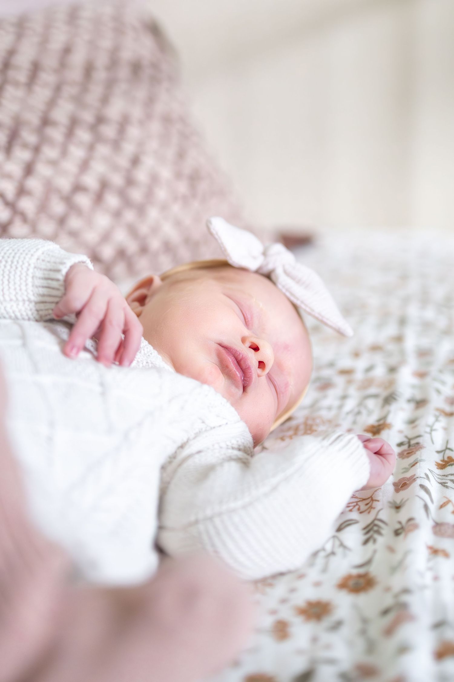 Sleeping baby in white sweater with bow accessory resting on floral bedding.