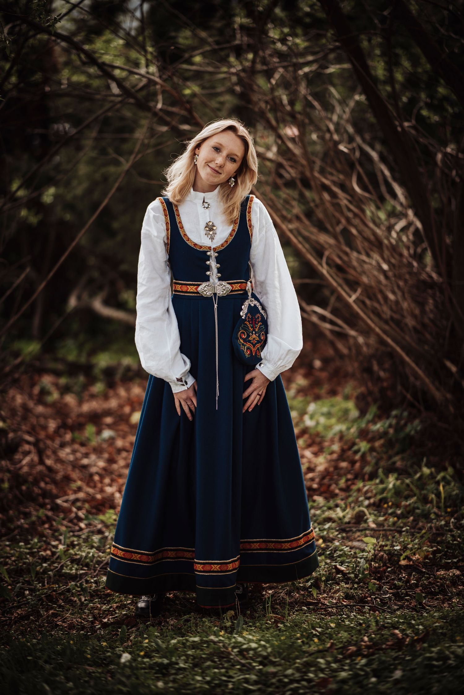 Traditional Norwegian bunad dress in navy blue with white blouse and decorative embroidery shown in outdoor woodland setting.
