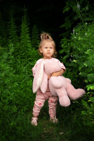 Small figure in pink outfit holding large plush bunny while standing among lush green ferns in garden setting.