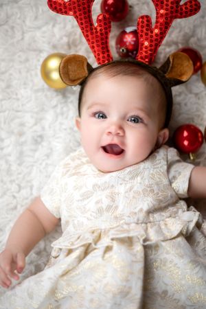 Holiday portrait with reindeer antler headband and lace dress surrounded by festive ornaments.