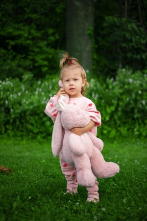 Pink-clad figure clutching stuffed bunny toy while standing on grass with green foliage background.