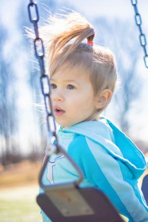 Bright sunlit portrait on playground swing wearing light blue hooded jacket.