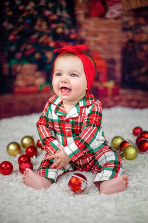 Festive holiday photo with plaid outfit and red bow sitting among Christmas ornaments.