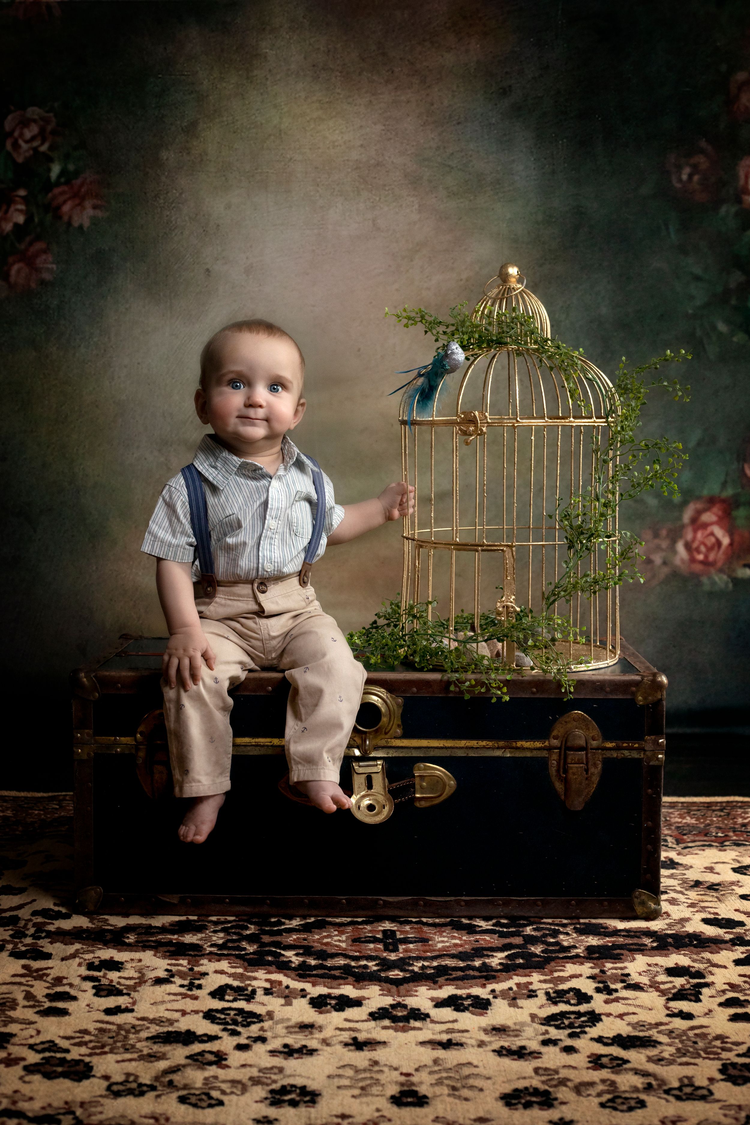 A baby in khaki overalls sits beside a decorative gold birdcage with greenery in a studio setting.