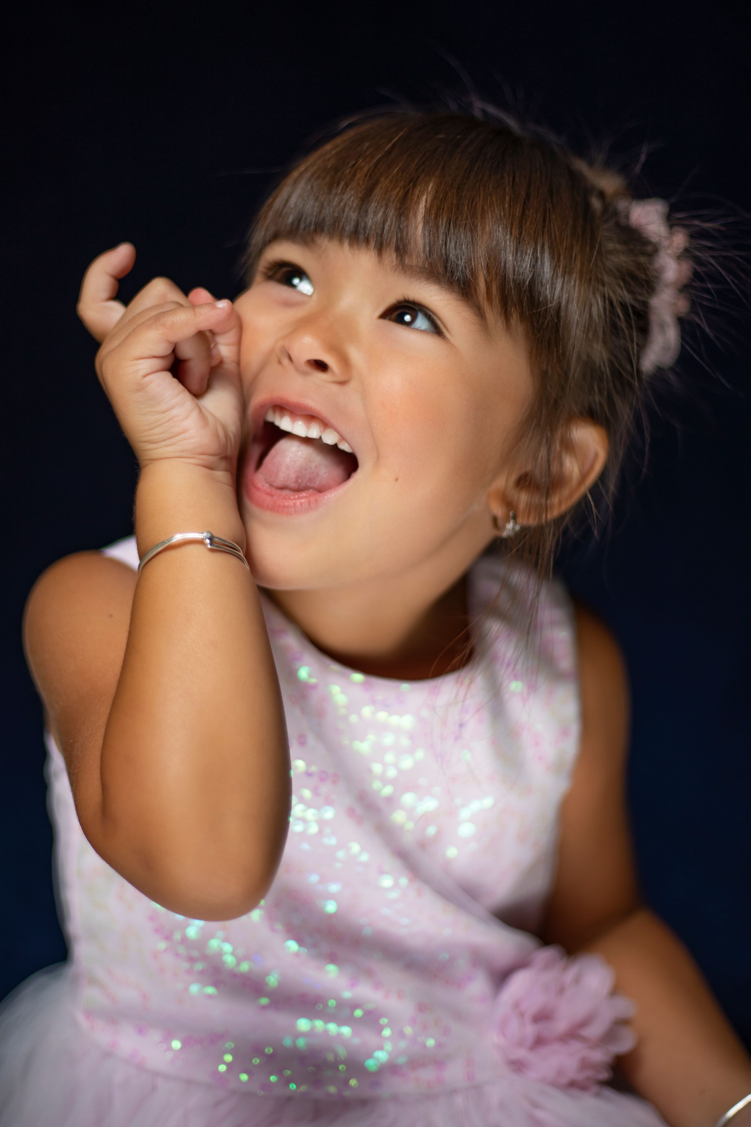 Young child in a sparkly pink dress making playful expressions against a dark background.