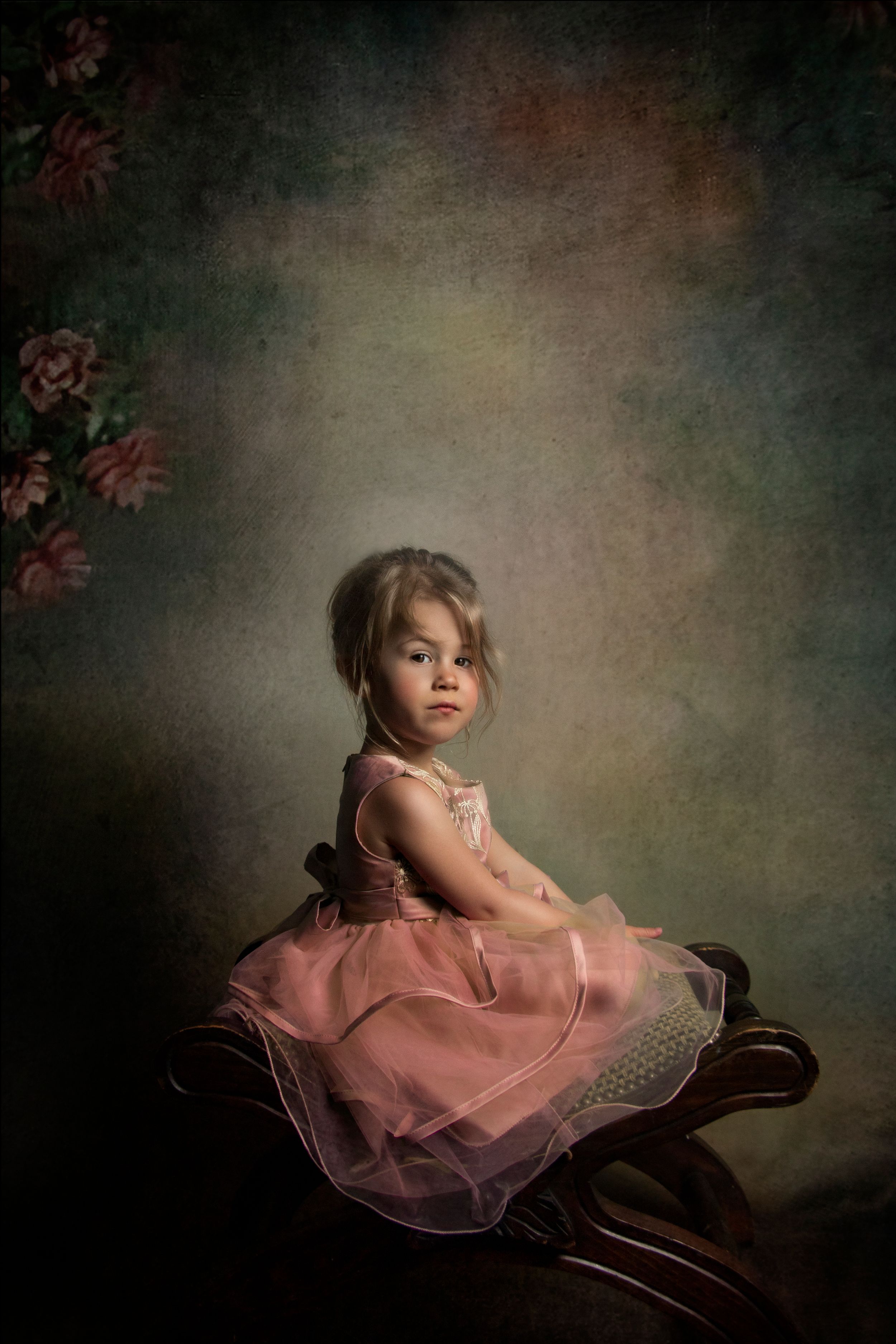 A vintage-style portrait of a small child in a pink tulle dress posed on an antique chair.