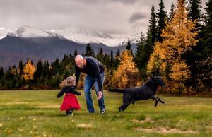 Family playing with black dog in autumn field with mountain backdrop and golden larches.