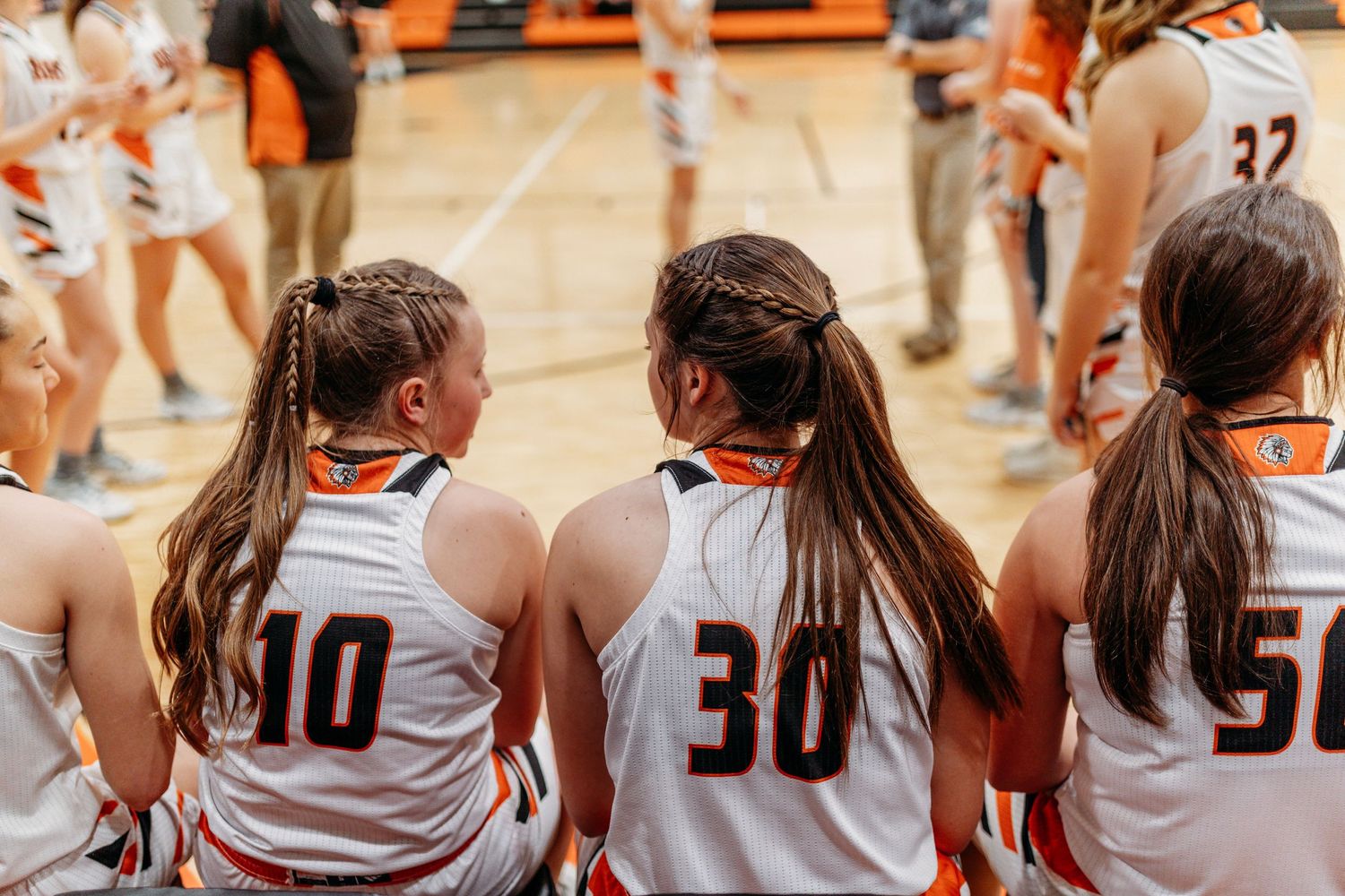 Girls basketball team huddles together on court wearing orange and white jerseys with numbers 10, 30, and 50.