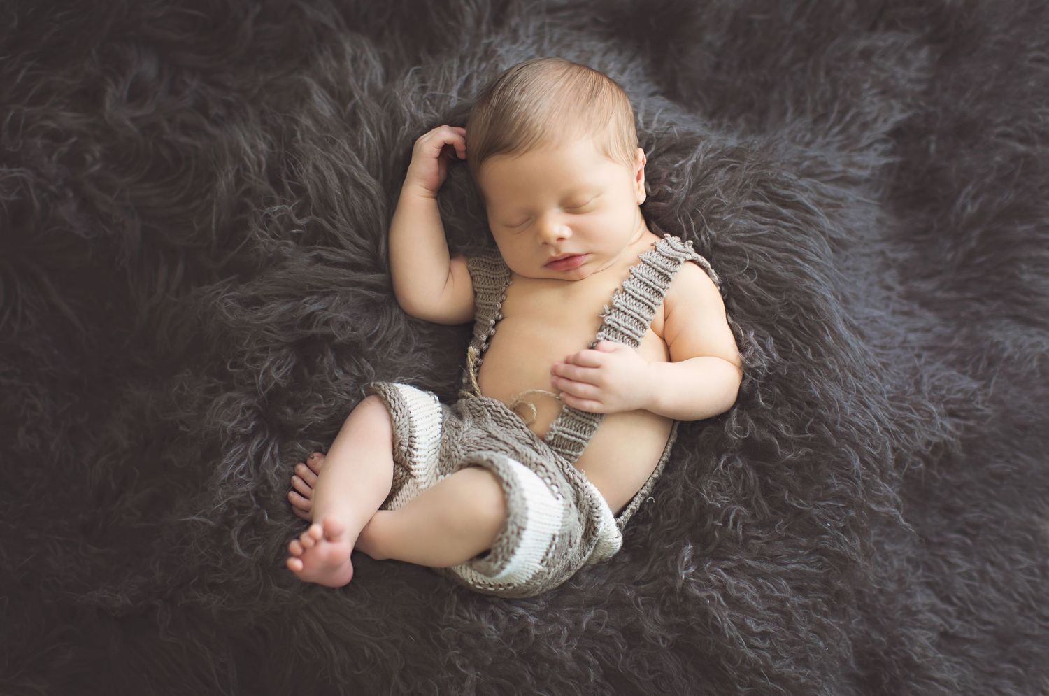 A sleeping newborn baby in gray clothing lies on a dark fluffy blanket.