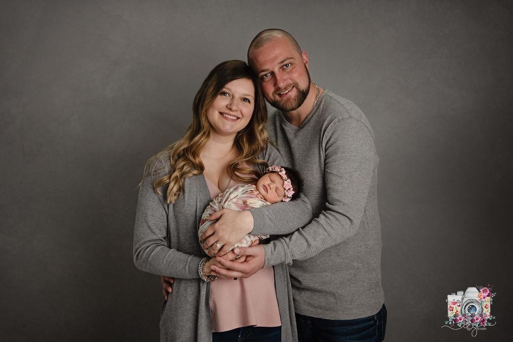 Family portrait against grey backdrop in matching sweaters holding their newborn baby.