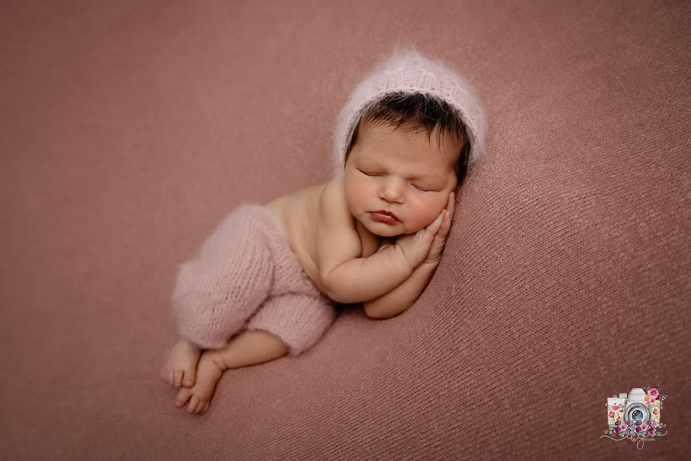 A sleeping newborn in soft pink clothing rests on a rose-colored backdrop.