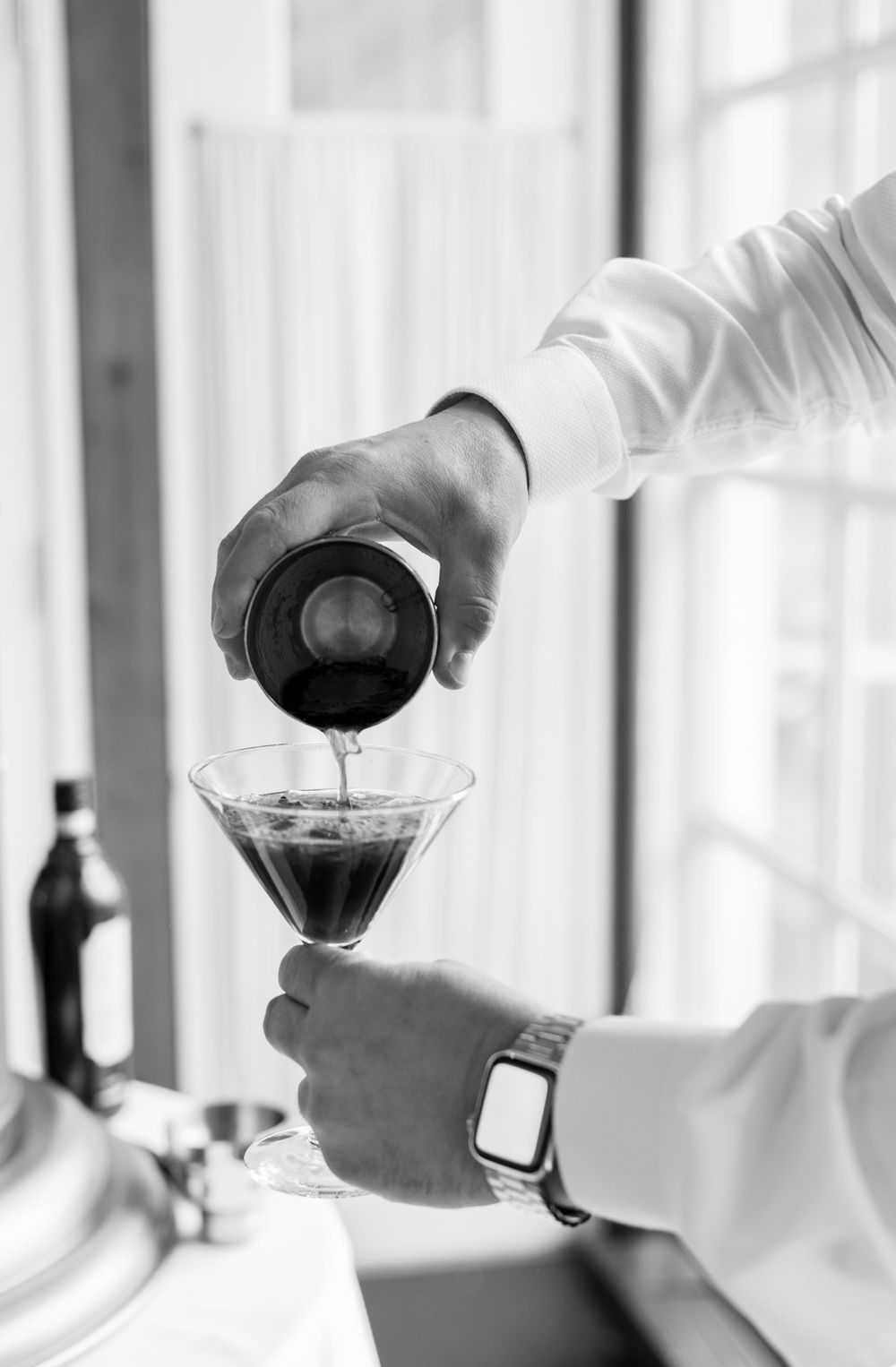 Close-up of hands pouring dark liquid into martini glass in black and white photography.