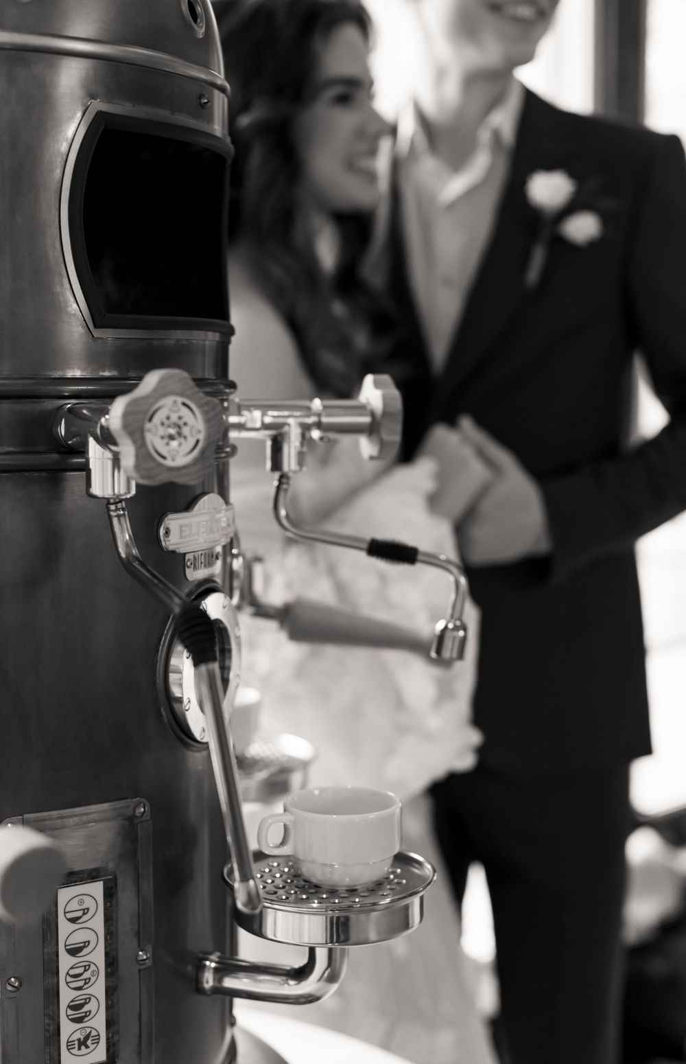 Vintage brass coffee machine in foreground with blurred wedding couple in background.