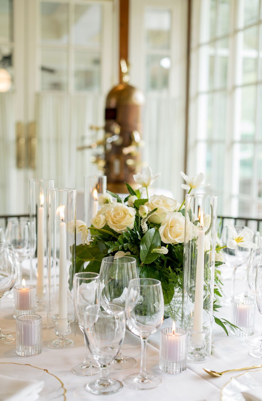 Elegant wedding table setting with white roses centerpiece, tall candles and wine glasses in a bright reception room.