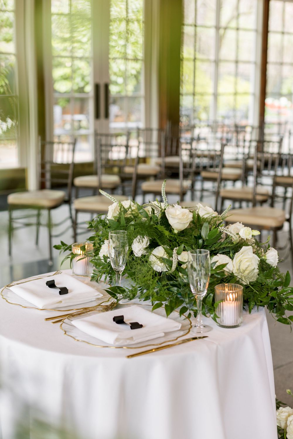 Elegant wedding table setting with white floral centerpiece and empty chairs in background.