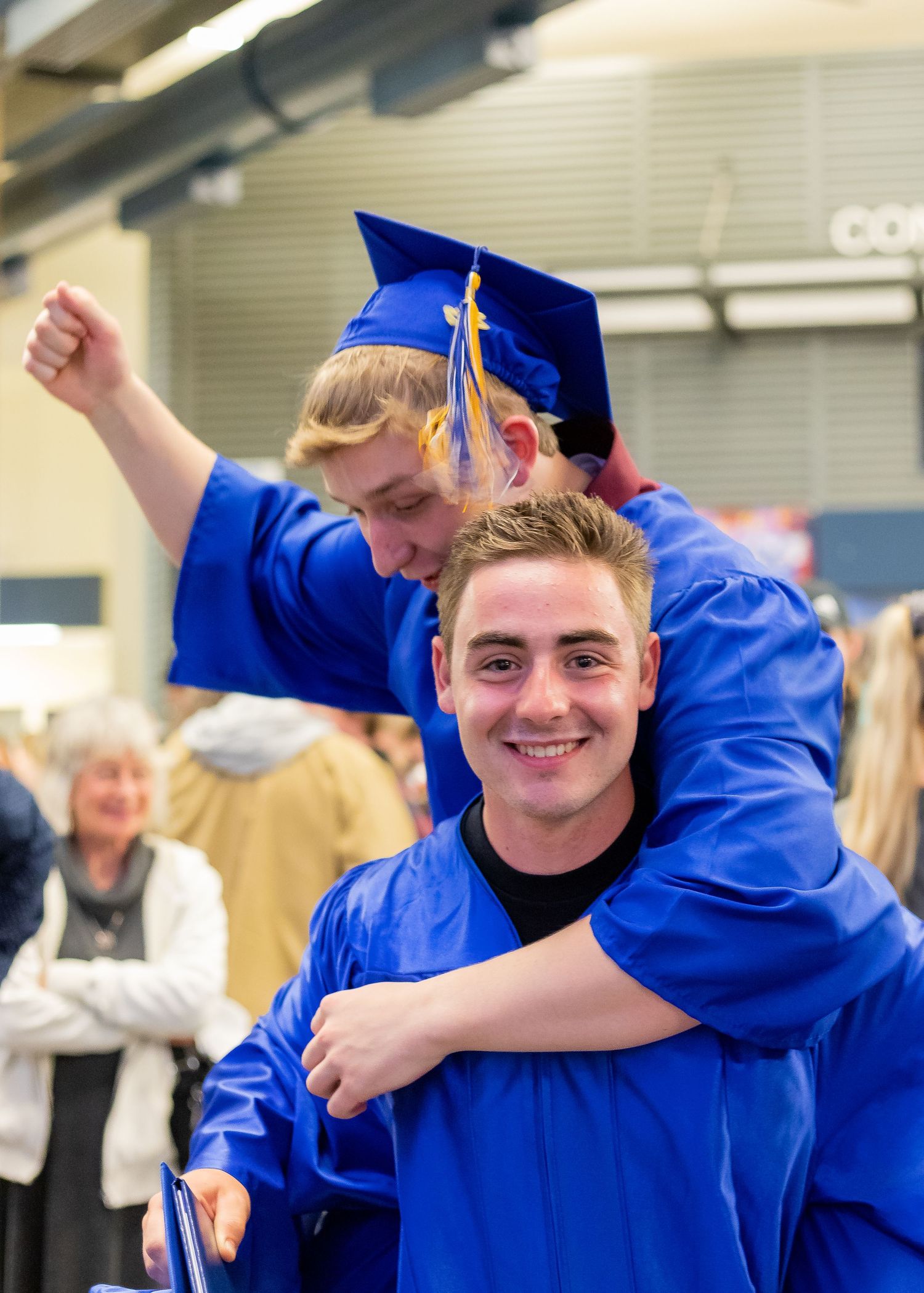 Two graduates in blue caps and gowns celebrate together with playful piggyback ride at graduation ceremony.