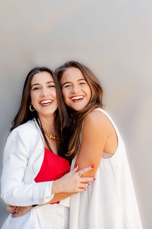 Two women in white and red outfits embracing and laughing against gray background.