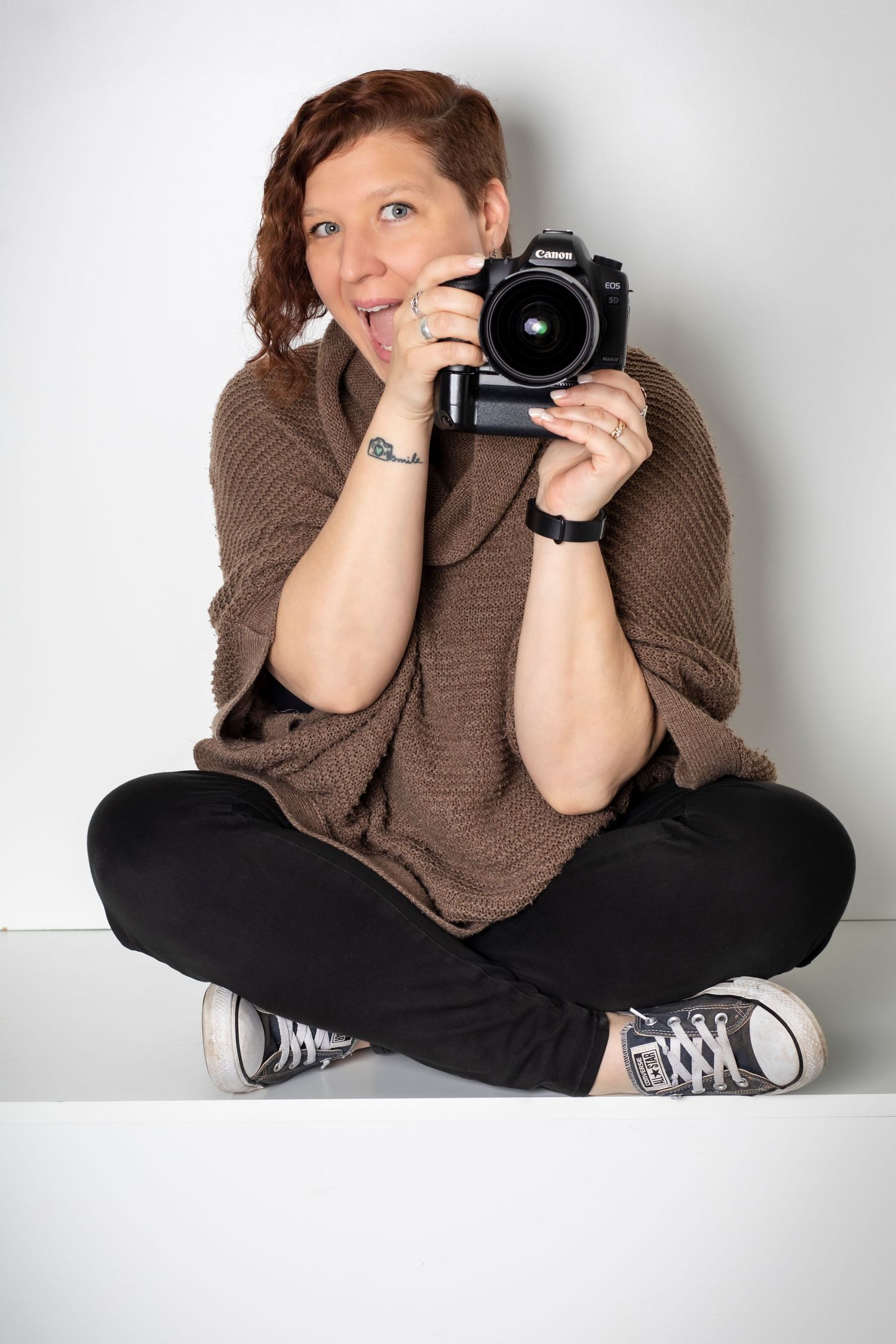 Casual photographer in brown sweater sitting cross-legged with camera against white background.