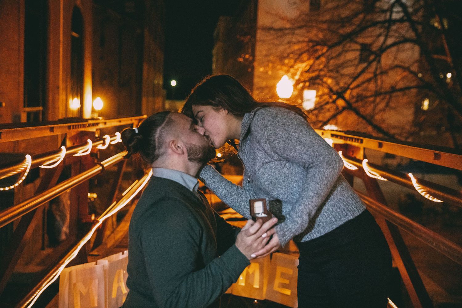Man proposes to woman on a warmly lit bridge at night as she leans in for a kiss.