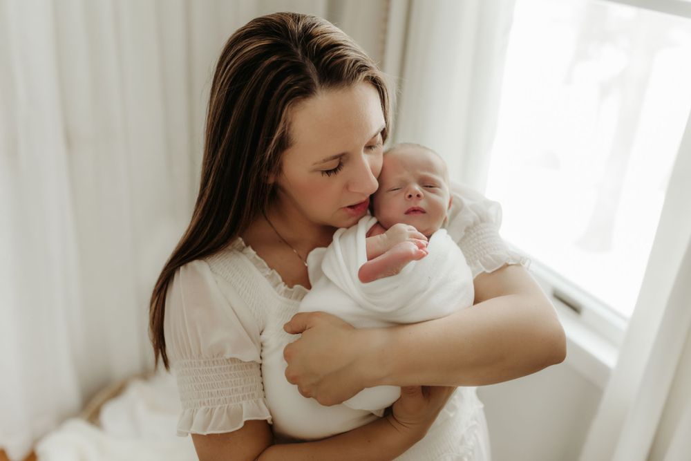 Tender moment by window with white-dressed woman holding swaddled infant.
