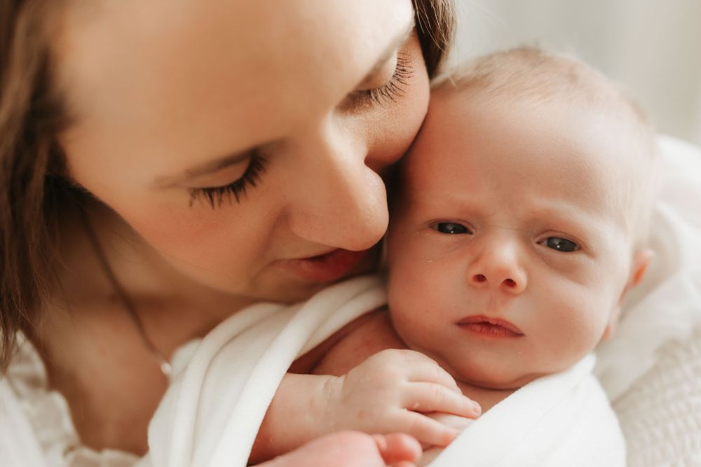 A tender moment captured between a parent and newborn baby in white clothing against a soft background.