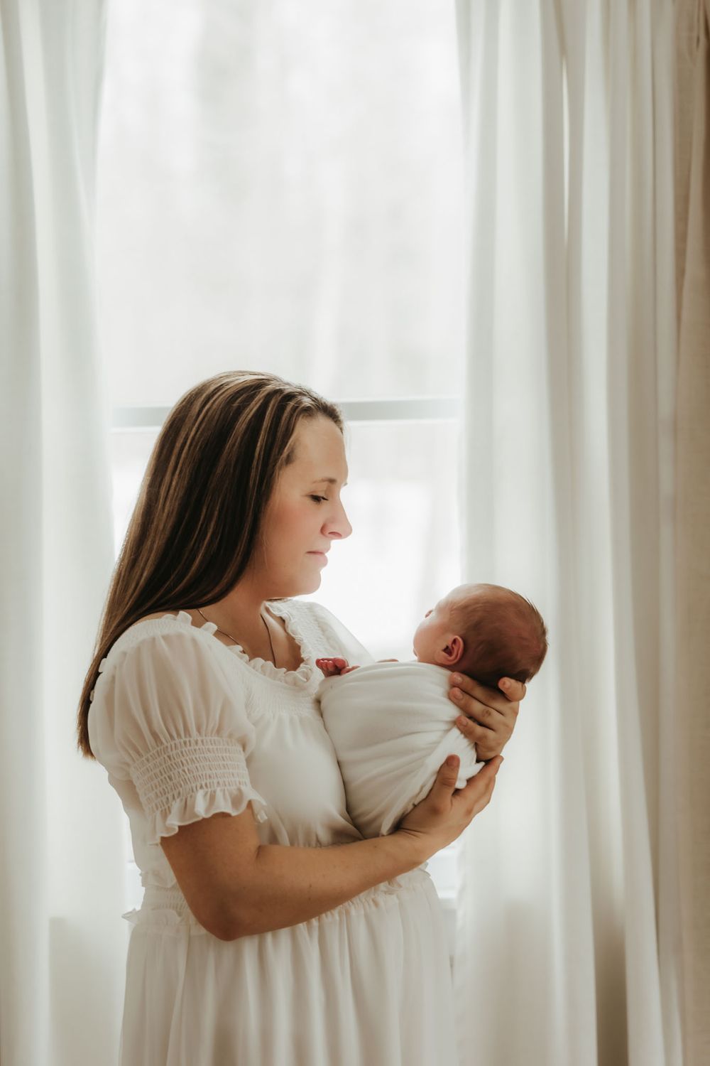 Backlit portrait of woman in white dress cradling newborn by sheer curtained window.