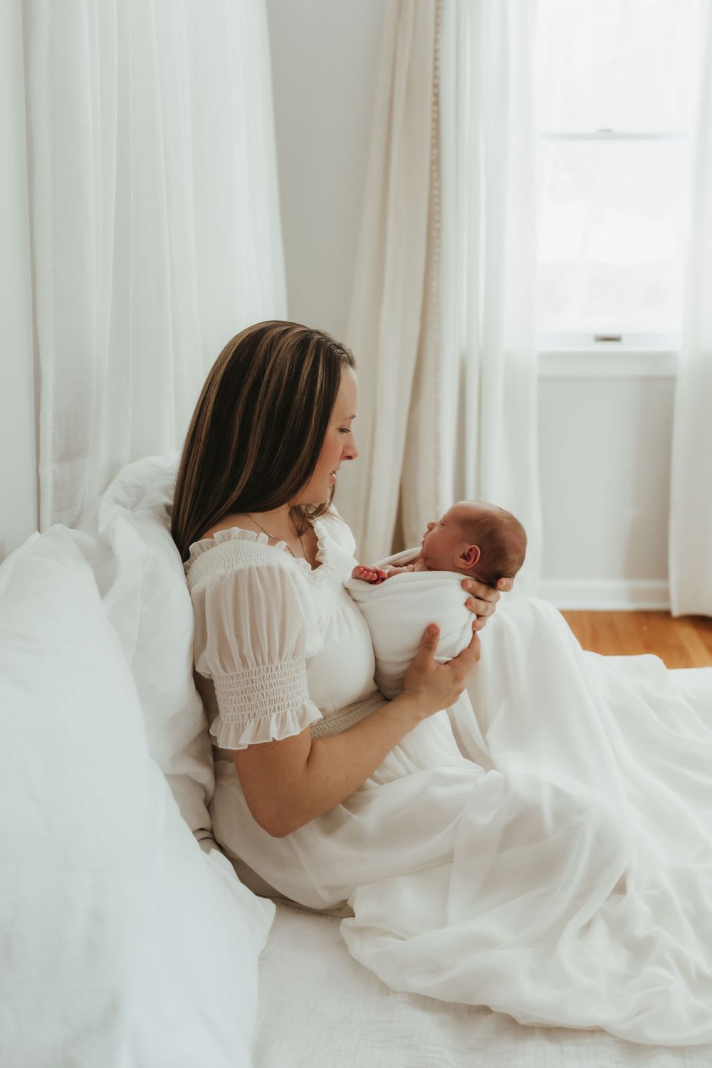 Parent in white dress sitting on bed while tenderly holding newborn baby.
