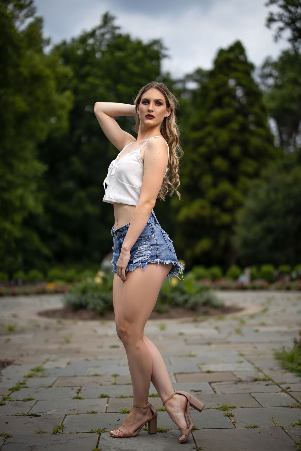 Model strikes pose in white tank top and distressed denim shorts on stone patio.