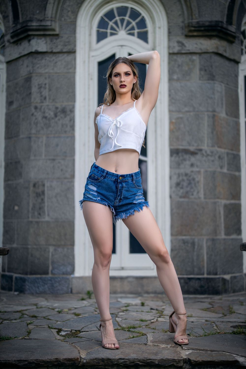 Fashion model posing in casual summer wear against historic stone building with arched doorway.